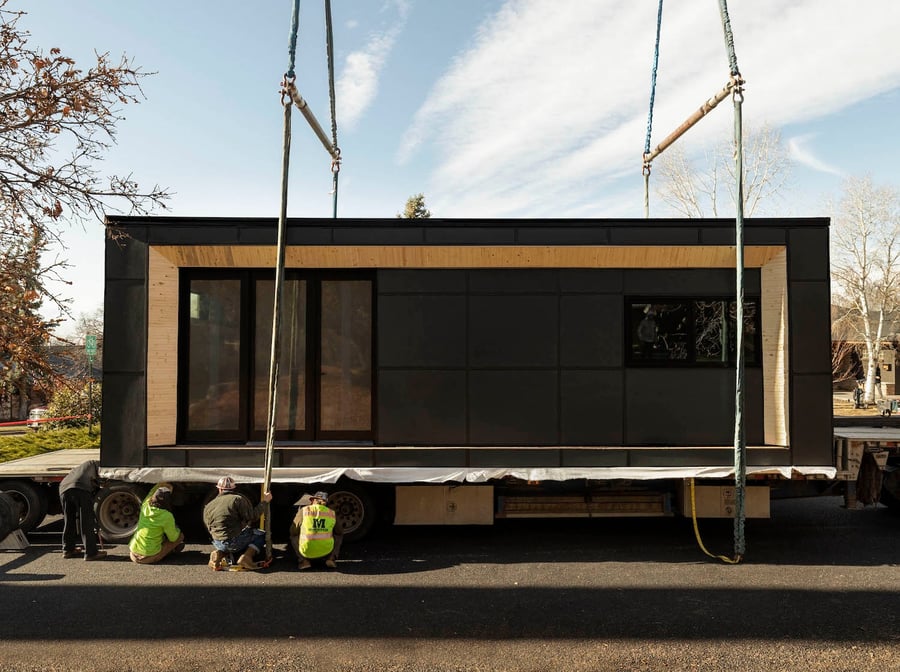 Small home sitting on a flatbed trailer with a crew hooking it to a crane