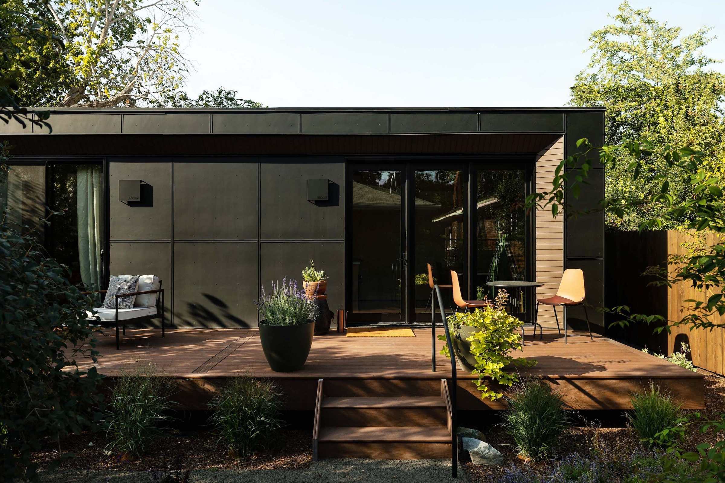 Dark, cozy exterior of a tiny house with sunlit porch in a backyard with trees