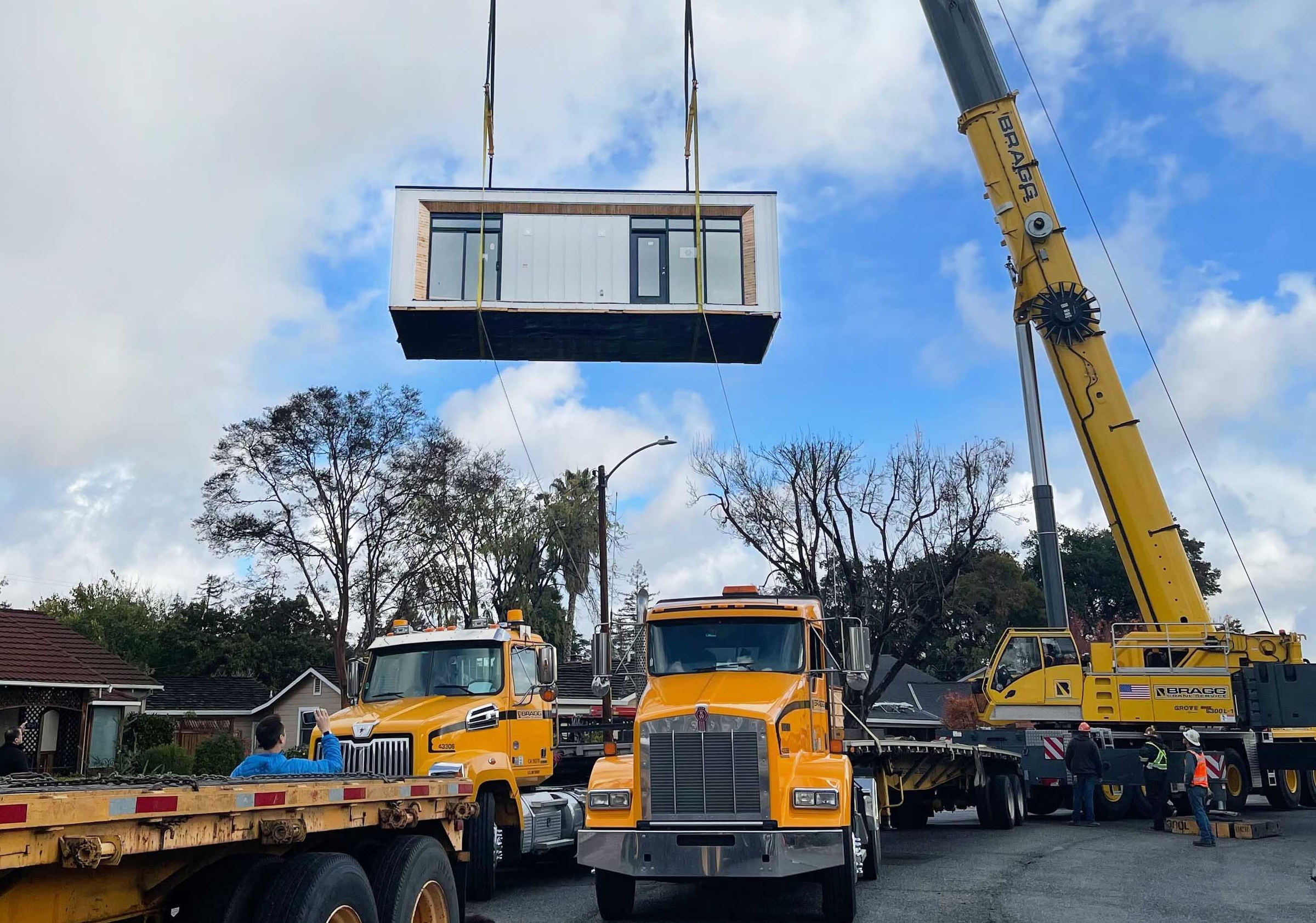 Modern, white accessory dwelling unit being craned over large semi trucks and a home